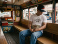 Man wearing a white Adobo graphic t-shirt and black adobo embroidered snapback hat sitting inside a colorful jeepney.